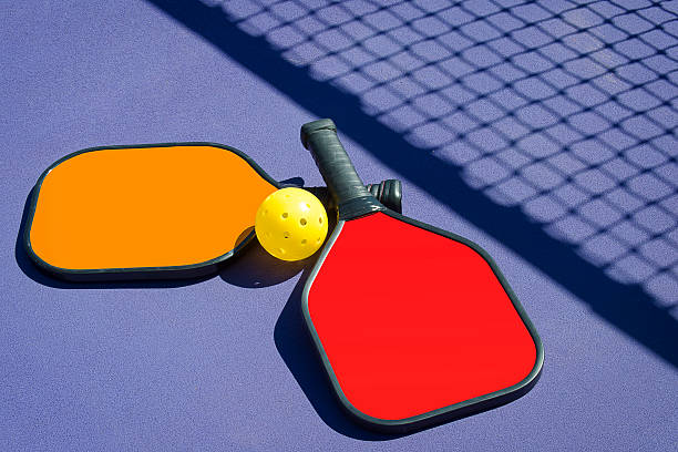 Stock photo of two (2) pickleball paddles and a yellow pickle ball lying on a blue playing court.